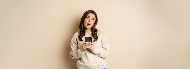 Online winkelen schattig meisje denken terwijl ze mobiele telefoon imaging smth dagdromen staande houdt