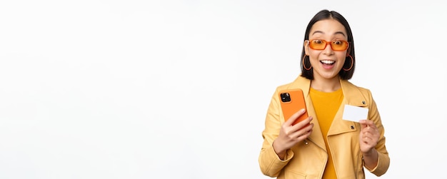 Online winkelen Stijlvol Aziatisch vrouwelijk model in zonnebril met creditcard en mobiele telefoon glimlachend gelukkig staande op witte achtergrond Ruimte kopiëren