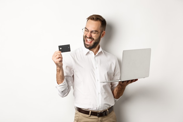 Online winkelen. Tevreden knappe man die naar creditcard kijkt na het bestellen van internet, met behulp van laptop, staande op een witte achtergrond