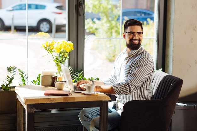 Online work. Joyful nice man smiling while working from the cafe on his laptop