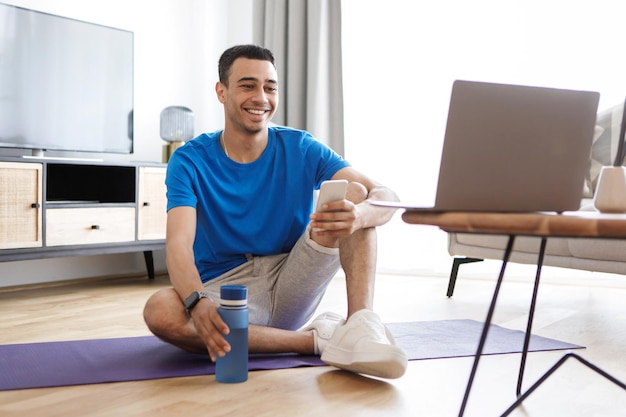 Online workout positive arab man exercising at home looking at\
laptop using smartphone having fitness class online