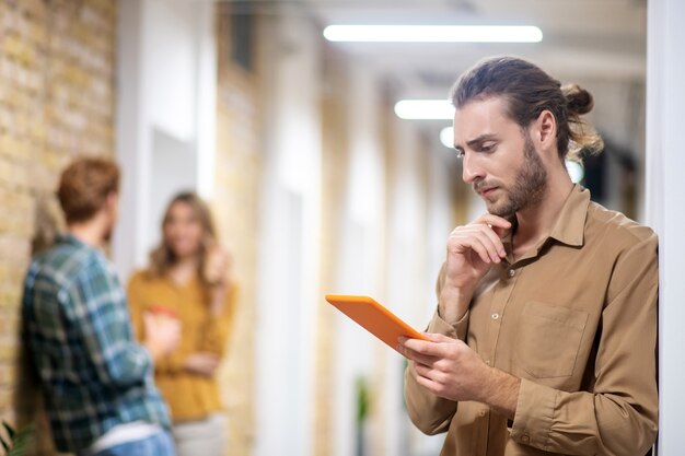 Online. Young bearded man standing in the corridor and holding a tablet in his hands