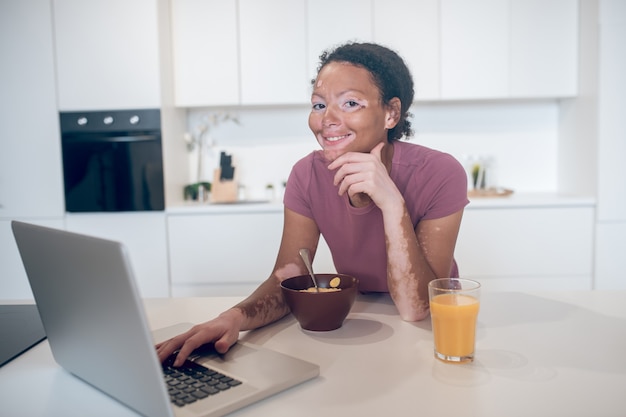 Online. Young mulatta in the kitchen watching something on internet and smiling