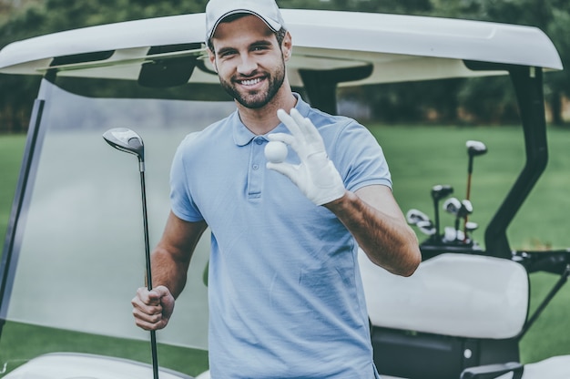 Foto solo la migliore attrezzatura da golf! bel giovane uomo sorridente che tiene in mano una pallina da golf e un autista mentre si trova vicino al carrello da golf e sul campo da golf