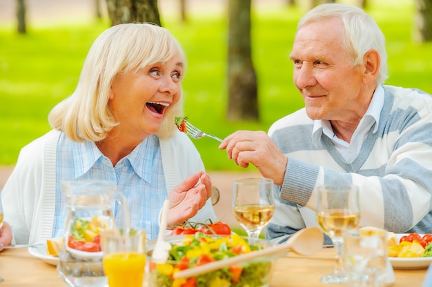 Only the best for our nearest. Senior man feeding his cheerful wife with fresh salad while both sitting at the dining table outdoors