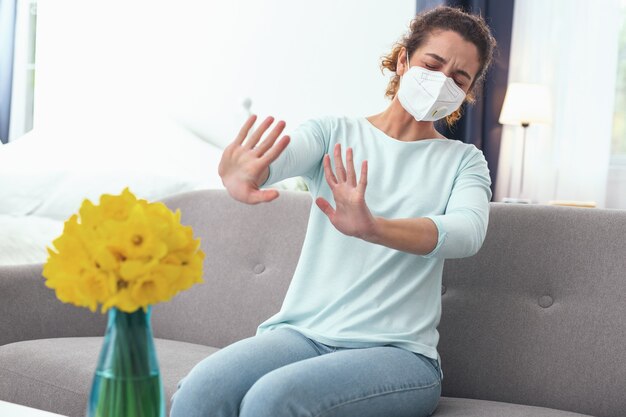 Onmogelijke geur. Jonge gekrulde vrouw die zich onwel voelt en overgevoelig is voor pas gebloeide lentebloemen terwijl ze een ademhalingsmasker draagt dat haar niet helpt