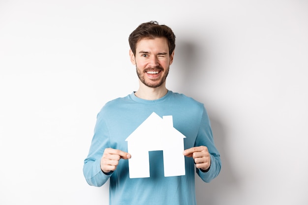 Onroerend goed en verzekeringsconcept. Gelukkig jongeman knipogen en glimlachen, papier huis knipsel tonen, staande op een witte achtergrond.