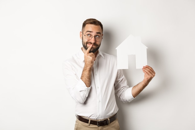 Onroerend goed. Man denkt tijdens het zoeken naar een appartement, houdt een papieren huismodel vast, staat op een witte achtergrond