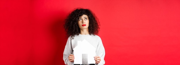 Onroerend goed trieste vrouw die droomt van het kopen van een appartement met een knipsel van een papieren huis en het opzoeken van nood