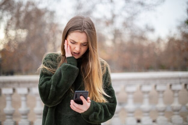 Onrustige jonge vrouw die voorhoofd wrijft en moderne smartphone bekijkt terwijl status in de herfstpark