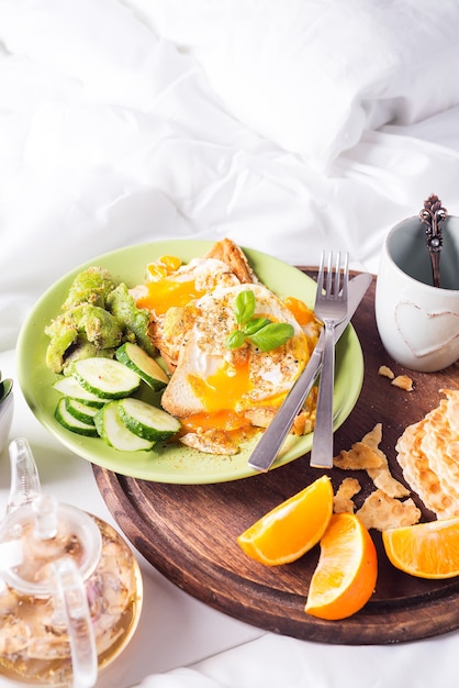 Foto ontbijt eieren op een witte plaat met halve avocado, komkommers op een houten plaat op bed