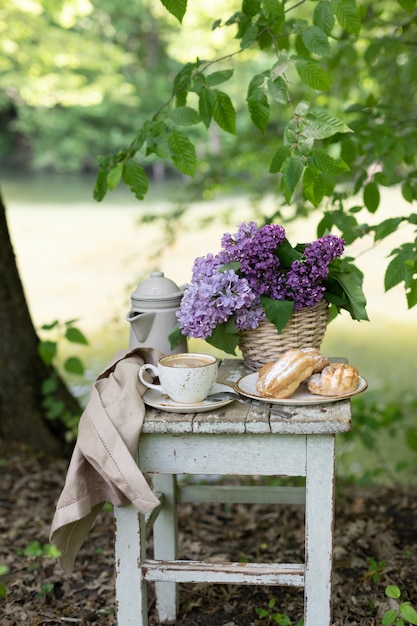 Ontbijt in de tuin: eclairs, kopje koffie, koffiepot, lila bloemen in een mandje.