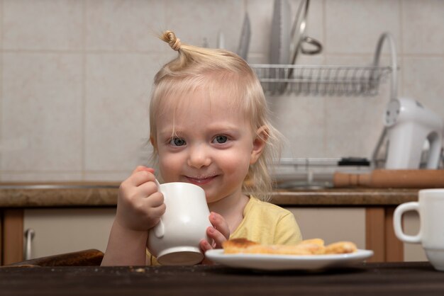 Ontbijt met kind. Het meisje drinkt een melk. Leuk blondekind met kop in handen in keuken.