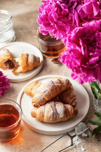 Ontbijt met twee platen van croissants thee suikerpot en pioenrozen bloemen op bruine achtergrond