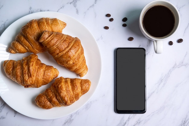 Ontbijt met verse croissants en kopje zwarte koffie, lege scherm slimme telefoon, bovenaanzicht