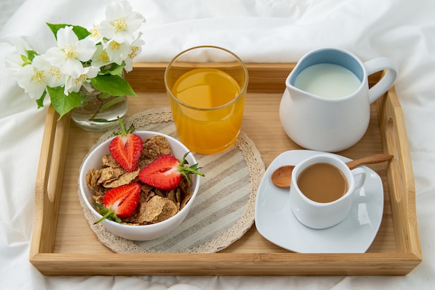 Foto ontbijt op bed. houten dienblad met koffie, jus d'orange, aardbeien en muesli. sieraden met fijne witte bloemen.