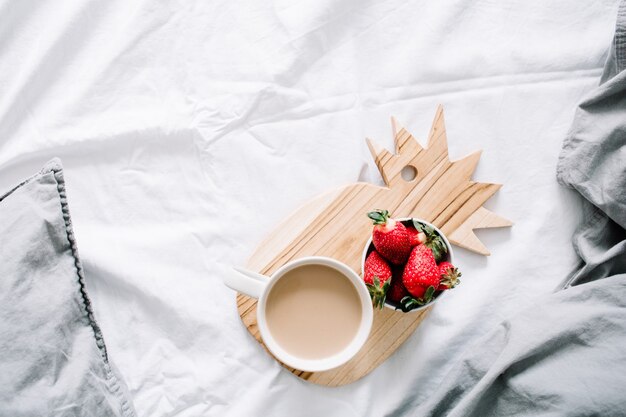 Ontbijt op bed met koffiemok en aardbei. Helder grijs beddengoed. Plat lag, bovenaanzicht.