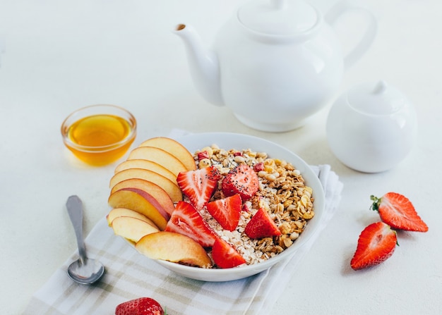 Ontbijt pap havermout met granen muesli aardbeien perziken honing in een witte plaat op de achtergrond van een witte theepot en kommen met honing op een witte plaat