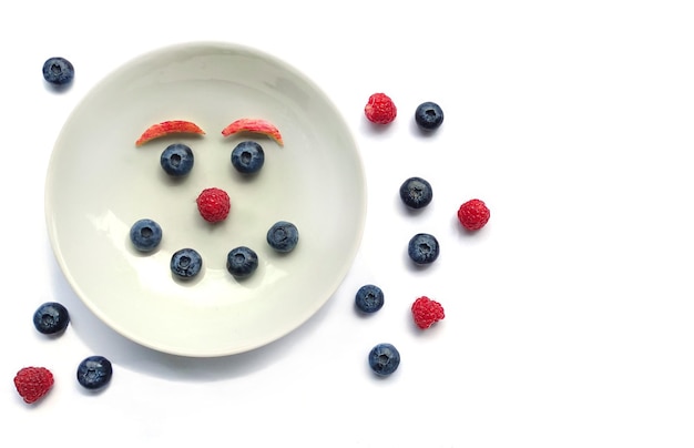 Ontbijt, snack voor kinderen. Grappig gezicht gemaakt van bosbessen en frambozen op een wit bord