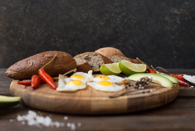 Ontbijt van gebakken eieren, brood, avocado en verschillende kruiden op een houten bord.