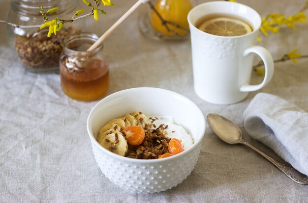 Ontbijt van muesli met yoghurt en thee en forsythia bloemen op een linnen tafellaken. Rustieke stijl.