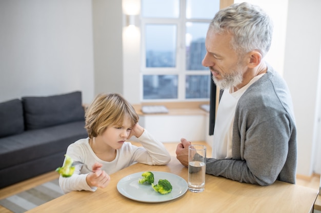 Ontbijt. Volwassen man probeert zijn zoon gezond te laten eten terwijl de jongen er ongelukkig uitziet