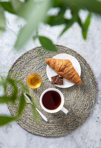 Ontbijtconcept met een kopje thee, honing en een croissant op een marmeren tafel en groene takken close-up bovenaanzicht verticale oriëntatie