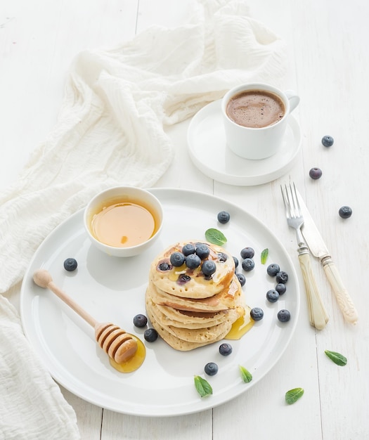 Ontbijtset Bosbessenpannenkoekjes met verse bessen, honing, muntblaadjes en kopje koffie op een witte houten achtergrond