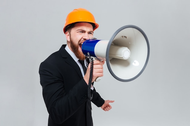 Ontevreden bebaarde ingenieur in zwart pak en oranje helm die in megafoon schreeuwt en opzij kijkt.