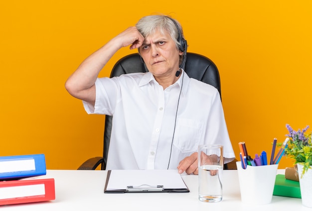 Ontevreden blanke vrouwelijke callcenter-operator op koptelefoon zittend aan bureau met kantoorgereedschap hand op voorhoofd geïsoleerd op oranje muur