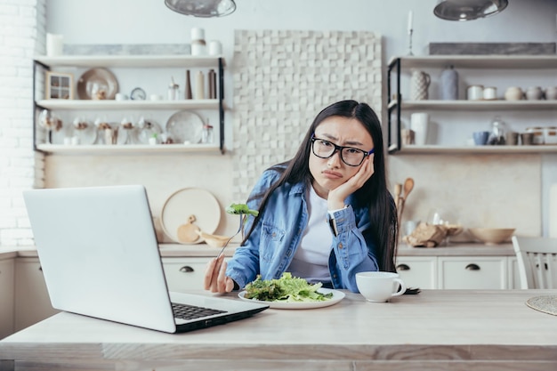 Ontevreden en moe van het op dieet zijn van een jonge aziatische vrouw, hij zit aan de tafel in de keuken