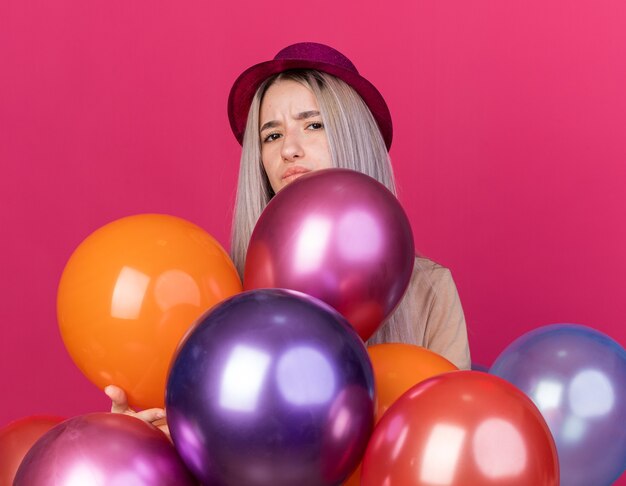 Ontevreden jong mooi meisje met feestmuts met beugels achter ballonnen geïsoleerd op roze muur