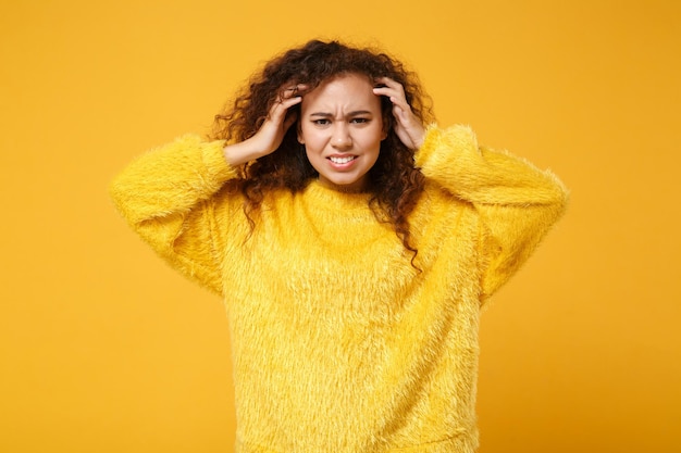 Ontevreden jonge Afro-Amerikaanse meisje in bont trui poseren geïsoleerd op geel oranje achtergrond studio portret. Mensen oprechte emoties levensstijl concept. Bespotten kopie ruimte. Handen op het hoofd zetten.