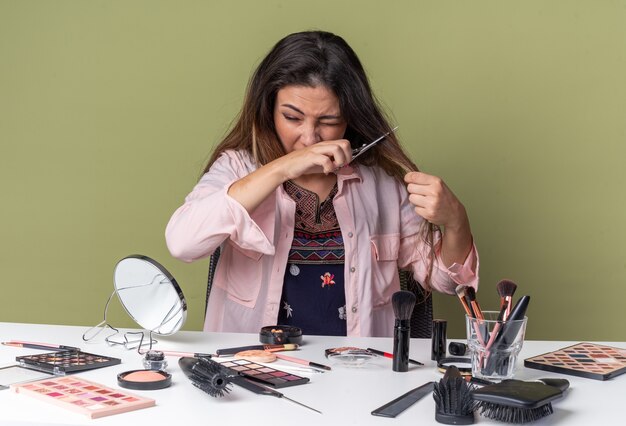 Foto ontevreden jonge brunette meisje zit aan tafel met make-up tools die haar haar knippen met een schaar geïsoleerd op olijfgroene muur met kopieerruimte