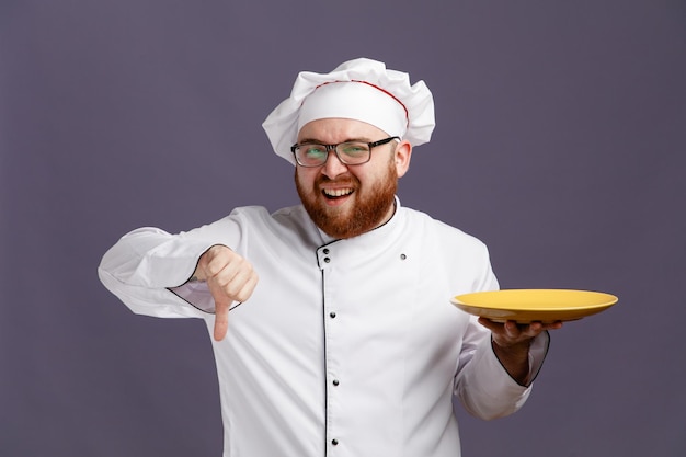 Ontevreden jonge chef-kok met een uniforme bril en een pet met een lege plaat die naar een camera kijkt met duim omlaag geïsoleerd op een paarse achtergrond