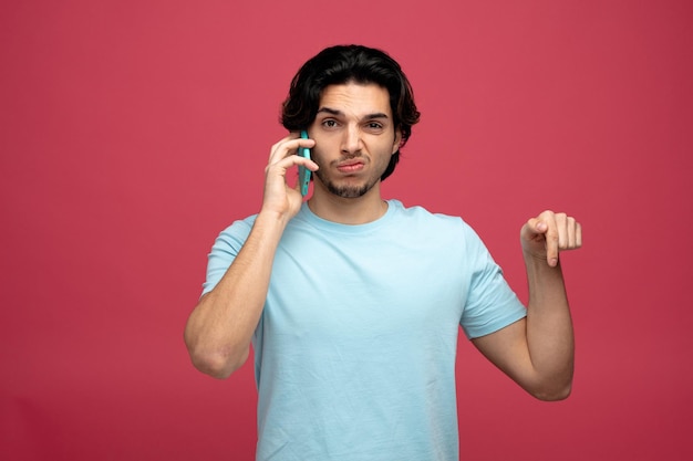 ontevreden jonge knappe man die naar de camera kijkt en naar beneden wijst terwijl hij aan de telefoon praat geïsoleerd op rode achtergrond