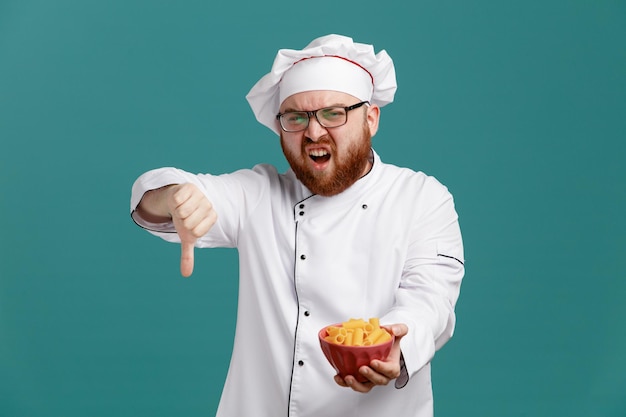 Ontevreden jonge mannelijke chef-kok met een uniforme bril en een pet met een kom macaroni-pasta die naar een camera kijkt met duim omlaag geïsoleerd op blauwe achtergrond