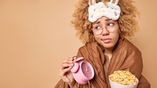 Ontevreden jonge vrouw wordt vroeg in de ochtend wakker en gaat ontbijten houdt wekker en kom cornflakes geconcentreerd weg poses tegen bruine achtergrond mock-up ruimte voor uw tekst