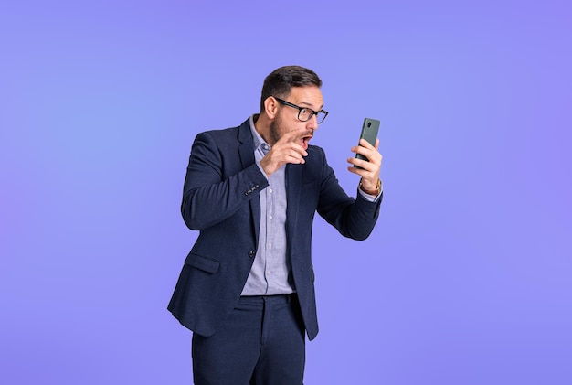 Ontevreden jonge zakenman gekleed in formele kleding die e-mails leest op mobiele telefoon Bezorgde mannelijke professionele ondernemer die mobiele telefoon gebruikt terwijl hij geïsoleerd op blauwe achtergrond staat