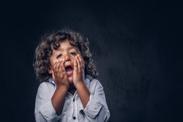 Ontevreden schooljongen met bruin krullend haar gekleed in een witte, houdt handen op het gezicht in een studio. Geïsoleerd op de donkere gestructureerde achtergrond.