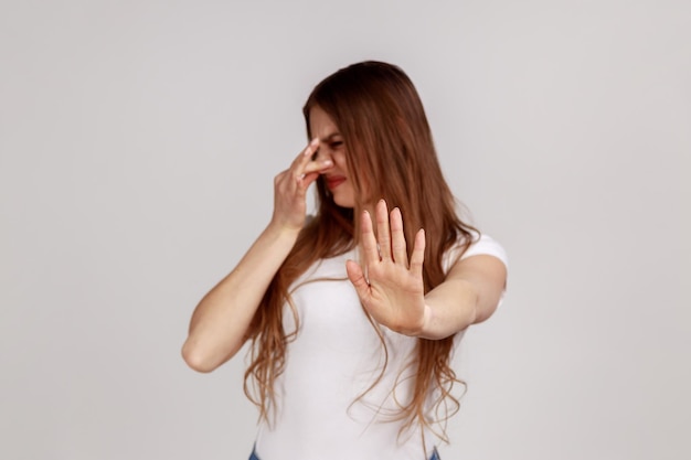 Ontevreden vrouw grimassen met walging adem inhouden neus knijpen met vingers om slechte geur te voorkomen en het tonen van stop gebaar dragen witte Tshirt Indoor studio opname geïsoleerd op grijze achtergrond