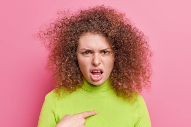 Ontevreden vrouw met krullend haar kijkt met boze verontwaardigde uitdrukking wijst naar zichzelf vraagt waarom geef je mij de schuld houdt mond open gekleed in casual groene poloneck geïsoleerd over roze muur