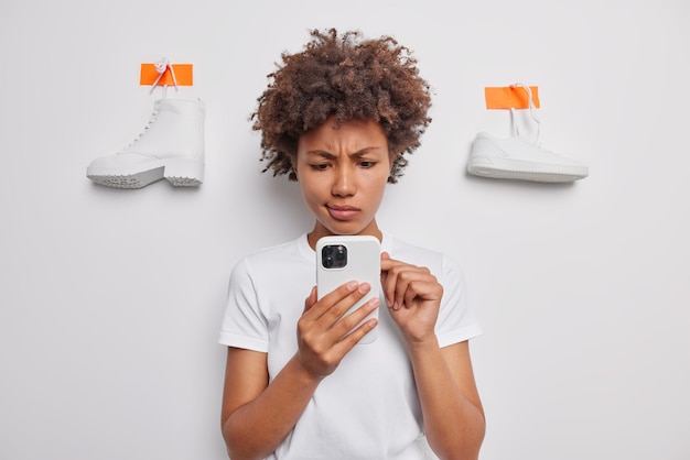 Ontevreden vrouw scrollt nieuwsfeed via smartphone fronst gezicht voelt ontevreden gekleed in casual t-shirt geïsoleerd over witte achtergrond met gepleisterde laars en sneaker aan muur Online winkelen