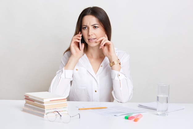 Ontevreden vrouwelijke professor praat op mobiele telefoon, draagt wit overhemd, bespreekt iets met verontwaardigde uitdrukking, heeft bril, stapel boeken, glas water op bureau. Mensen, zaken, carrière