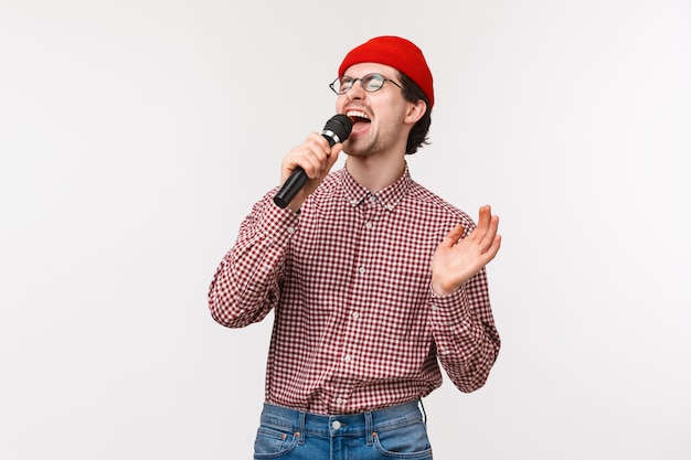 Ontroostbare kerel die romantisch lied in karaoke zingt met hartstochtelijk gevoel, ogen sluit en in microfoon schreeuwt, staand in rode muts, bril en geruit hemd, op een witte muur