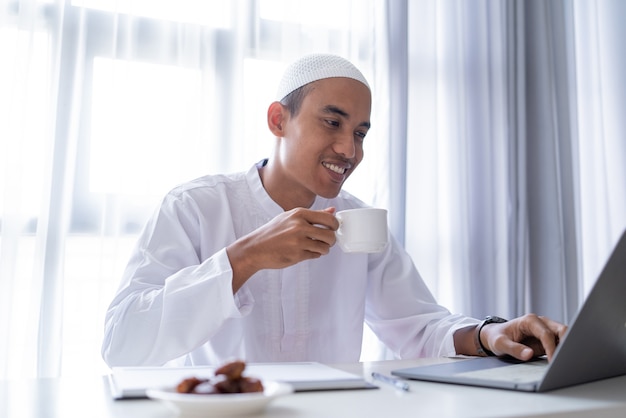 Ontspan Aziatische musim man met een kopje koffie terwijl hij thuis werkt met behulp van een laptop