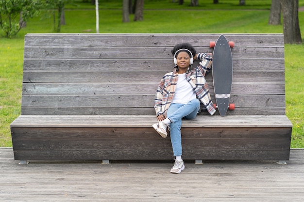 Ontspannen Afrikaanse vrouwelijke skateboarder chill luister muziek met koptelefoon en longboard in stedelijke ruimte