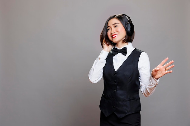 Ontspannen Aziatische serveerster luisteren naar muziek in koptelefoon. Glimlachende vrolijke restaurantvrouw-werknemer die uniform draagt en geniet van liedjes in draadloze oortelefoons terwijl ze poseert in de studio
