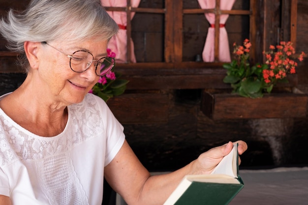 Ontspannen blanke senior vrouw in het wit met een bril die buiten het huis zit en een boek leest