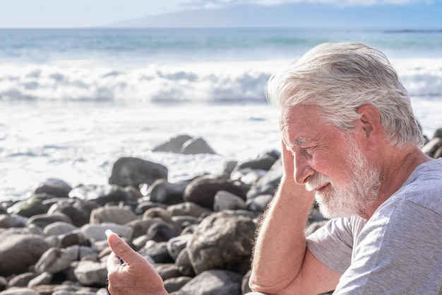 Ontspannen en lachende volwassen bebaarde senior man zittend op het rotsstrand met behulp van mobiele telefoon
